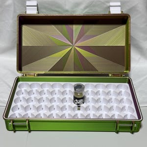 May include: Open green and gold metal case with a white interior tray. The case contains a small glass bottle with a black and white design. The case has a gold interior with a starburst design.