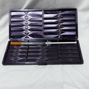 May include: A dark purple, open case with a geometric pattern, containing a syringe with an orange tip. The case is divided into multiple compartments. The background is a white fabric.