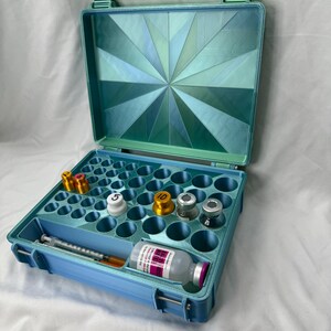 May include: A light blue storage case with a geometric starburst pattern on the interior lid. The base features multiple circular slots containing small metal vials, some marked with numbers, alongside a syringe and a clear bottle with a pink label.