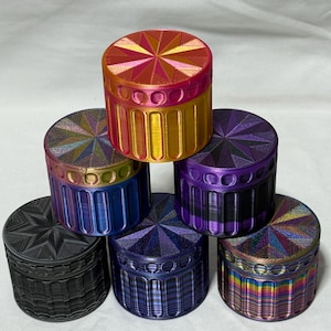May include: A collection of colorful, cylindrical containers with textured lids. The containers are stacked, showcasing a variety of colors including gold, purple, blue, and black. The lids feature starburst and geometric patterns.