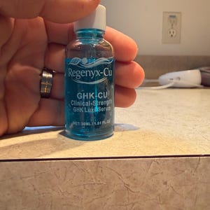 May include: A small, clear glass bottle with a blue liquid and a white dropper. The bottle has a white cap and is labeled "Regenyx-Cu GHK-CU Clinical-Strength GHK Luxe Serum". The bottle contains 1.01 fl oz (30ml).