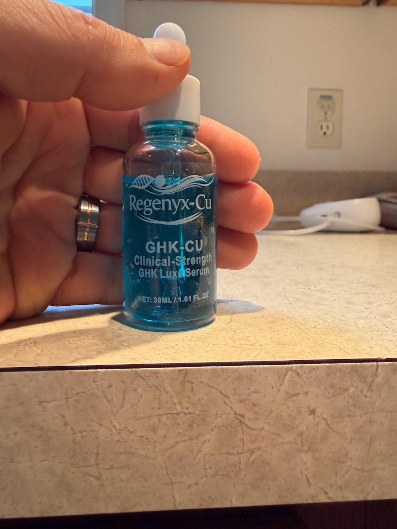 May include: A small, clear glass bottle with a blue liquid and a white dropper. The bottle has a white cap and is labeled "Regenyx-Cu GHK-CU Clinical-Strength GHK Luxe Serum". The bottle contains 1.01 fl oz (30ml).
