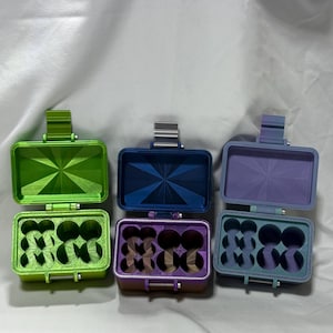 May include: Three open, rectangular containers with hinged lids. The containers are in green, blue, and purple. Each container has a geometric design on the lid and holds several small, round objects. The containers are on a white surface.