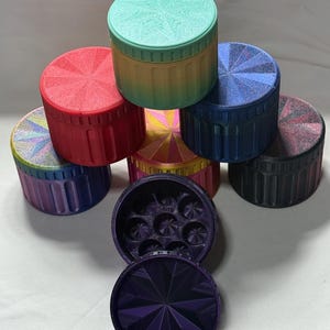 May include: A collection of colorful, cylindrical containers with textured lids. The containers are in various colors, including red, blue, green, and purple. One container is open, revealing a compartmented interior. The lids have a starburst design.