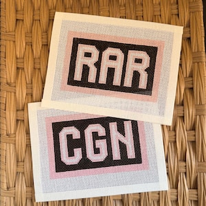 May include: Two rectangular needlepoint canvases featuring block lettering. The top canvas displays the letters RAR in light pink against a black background, framed by a light pink border. The bottom canvas shows CGN in light pink on black.
