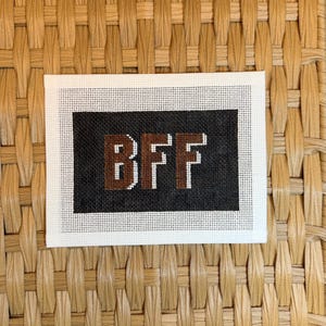 May include: A rectangular needlepoint canvas featuring a black background with the letters BFF stitched in brown with a white outline. The canvas has a white border and a visible grid pattern throughout the design, showcasing the needlepoint work.