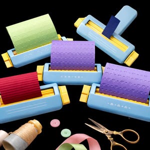 May include: A collection of blue and yellow paper embossing machines with various colors of embossed paper. The paper has patterns of stars, hearts, and waves. Also included are scissors, buttons, ribbon, and thread.