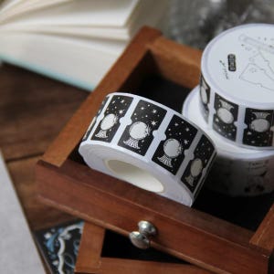 May include: Two rolls of adhesive paper tape featuring a repeating black and white design of crystal balls surrounded by stars. The tape is presented in a wooden box, with the top roll showing printed text and line art illustrations.