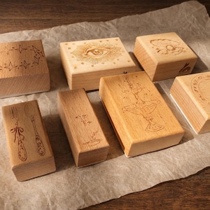 May include: A collection of seven light-coloured wooden rubber stamps featuring various engraved designs including an eye, a fountain, a fork and spoon, a shell, and circular patterns. Each stamp has a smooth, rectangular wooden block base.