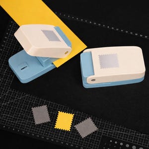 May include: Two light blue and cream-colored paper punches with a stamp-shaped design. One punch is open with a yellow paper strip inserted, while the other is closed. Three small stamp-shaped paper cutouts in gray and yellow are visible.