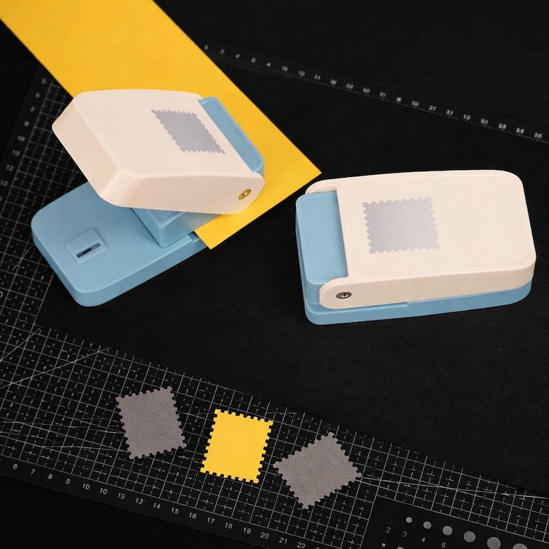 May include: Two light blue and cream-colored paper punches with a stamp-shaped design. One punch is open with a yellow paper strip inserted, while the other is closed. Three small stamp-shaped paper cutouts in gray and yellow are visible.