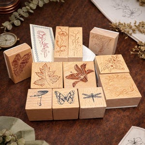 May include: A collection of wooden stamps with various botanical and insect designs. The stamps feature images of leaves, flowers, a mushroom, a butterfly, and a dragonfly. The stamps are arranged on a brown surface.