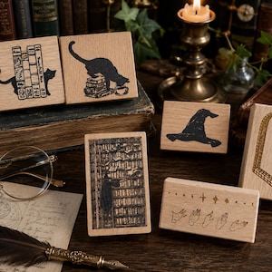 May include: A collection of six rectangular wooden rubber stamps featuring black ink designs including a cat with books, a cat on a stack of books, a pointed hat, a tall bookshelf, a stone archway, and a row of hands with celestial stars.