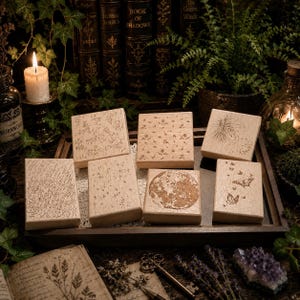 May include: A set of six rectangular wooden rubber stamps featuring engraved designs of rain, stars, a full moon, butterflies, wildflowers, and a dandelion seed head, arranged together on a dark wooden tray.