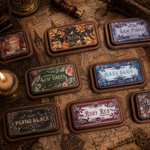 May include: A collection of rectangular ink pads in copper-colored metal cases, each featuring a unique floral and bird illustration with labels including Yellow, Lilac Purple, Leaf Green, Haze Blue, Piano Black, and Ruby Red in decorative fonts.