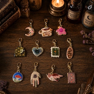May include: A collection of eleven gold-toned metal charms featuring various enamel designs including a crescent moon, a mug labelled Witch's Brew, a pink flower, a cat, a mushroom, a snake, a hand with an eye, a bird, and a seed packet.