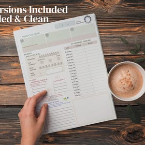 以下が含まれることがあります： 「2 Versions Included Guided & Clean」と「Today's Slogan」の文字が入った白いプランナーが木製の表面に開かれています。 手がプランナーを指しています。 コーヒーカップが近くにあります。 プランナーには、毎日のチェックインと成長タイムラインのセクションが含まれています。