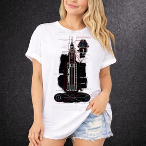 Peut inclure: T-shirt blanc avec un schéma détaillé du système de lancement spatial Artemis II. Le graphique comprend des composants étiquetés et un fond noir, avec le texte "ARTEMIS II" en haut. Le t-shirt est de style col rond.