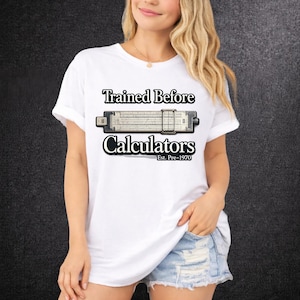 Peut inclure: T-shirt blanc avec l'inscription "Trained Before Calculators" et un graphique de règle à calcul. Le texte "Est. Pre-1970" est en dessous. La personne porte un short en jean bleu clair.