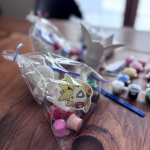 May include: A clear plastic bag filled with small white figurines, paint pots, and a paintbrush. A sticker of a cartoon character is attached to the bag. Several other bags and figurines are visible in the background, along with small paint pots.