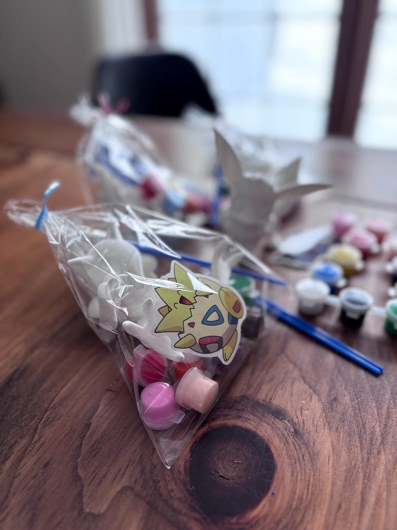 May include: A clear plastic bag filled with small white figurines, paint pots, and a paintbrush. A sticker of a cartoon character is attached to the bag. Several other bags and figurines are visible in the background, along with small paint pots.