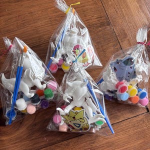 May include: Four clear plastic bags containing paint-your-own figurines. Each bag includes a white figurine, paint pots in various colors, and a blue paintbrush. The figurines are of different cartoon characters.
