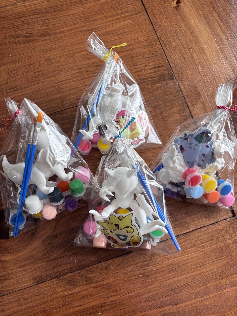 May include: Four clear plastic bags containing paint-your-own figurines. Each bag includes a white figurine, paint pots in various colors, and a blue paintbrush. The figurines are of different cartoon characters.