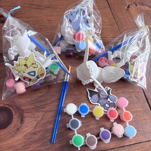 May include: Three clear cellophane bags tied with ribbon, each containing a white figurine, paint pots, and a paintbrush. The figurines are various cartoon characters. Paint pots are in a variety of colors, including pink, blue, and yellow.
