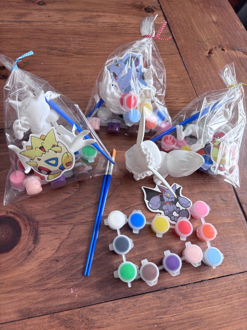 May include: Three clear cellophane bags tied with ribbon, each containing a white figurine, paint pots, and a paintbrush. The figurines are various cartoon characters. Paint pots are in a variety of colors, including pink, blue, and yellow.