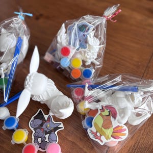 May include: Paint-your-own craft kits featuring white plaster figures, paint pots in various colors, and stickers. The kits are packaged in clear plastic bags, tied with blue and red ribbons. The kits include a variety of cartoon character stickers.