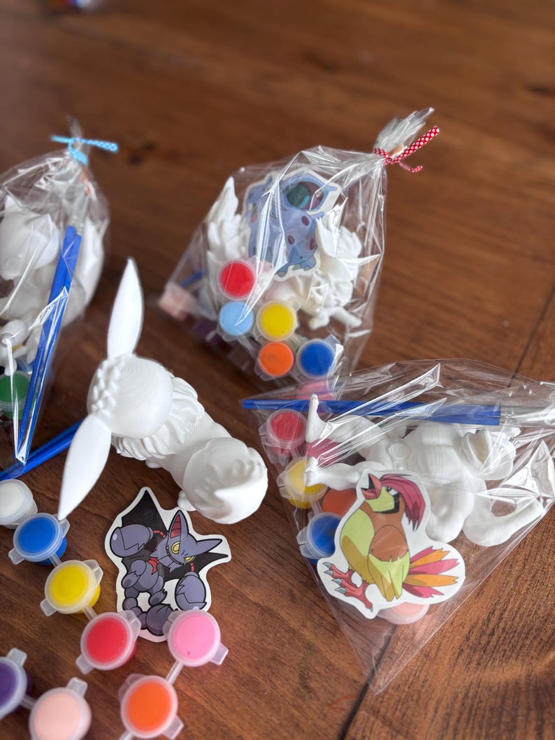 May include: Paint-your-own craft kits featuring white plaster figures, paint pots in various colors, and stickers. The kits are packaged in clear plastic bags, tied with blue and red ribbons. The kits include a variety of cartoon character stickers.