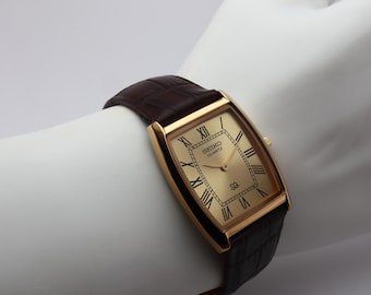 May include: A gold-toned Seiko Quartz watch with a rectangular face and Roman numeral hour markers. The watch has a brown leather band with a crocodile pattern. The watch is displayed on a white surface.