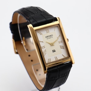Puede incluir: Reloj Seiko Quartz dorado con esfera rectangular y correa de cuero negro. La esfera es de color crema con n&uacute;meros romanos y agujas doradas. La correa tiene una textura con un patr&oacute;n de cocodrilo.