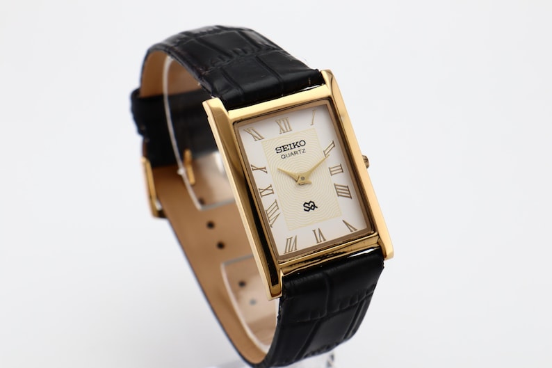 Puede incluir: Reloj Seiko Quartz dorado con esfera rectangular y correa de cuero negro. La esfera es de color crema con n&uacute;meros romanos y agujas doradas. La correa tiene una textura con un patr&oacute;n de cocodrilo.