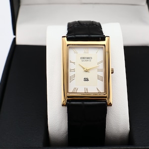 Puede incluir: Un reloj Seiko Quartz dorado con esfera rectangular y correa de cuero negro. La esfera del reloj es blanca con agujas doradas y n&uacute;meros romanos. El reloj se muestra en una caja negra con interior blanco.