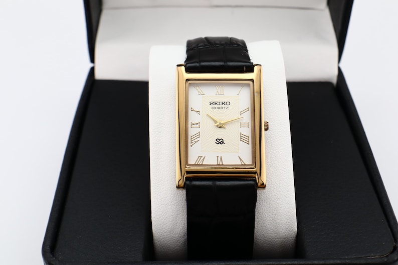 Puede incluir: Un reloj Seiko Quartz dorado con esfera rectangular y correa de cuero negro. La esfera del reloj es blanca con agujas doradas y n&uacute;meros romanos. El reloj se muestra en una caja negra con interior blanco.