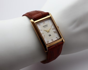May include: A gold-toned Seiko Quartz watch with a rectangular face and brown leather band. The watch face is cream-colored with Roman numerals and the Seiko logo. The band is a rich, textured brown.