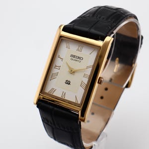 Puede incluir: Un reloj Seiko Quartz rectangular dorado con esfera crema y correa de cuero negro. La esfera del reloj presenta n&uacute;meros romanos y agujas doradas. La correa es de cuero negro con un patr&oacute;n texturizado.