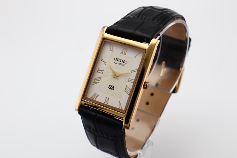 Puede incluir: Un reloj Seiko Quartz rectangular dorado con esfera crema y correa de cuero negro. La esfera del reloj presenta n&uacute;meros romanos y agujas doradas. La correa es de cuero negro con un patr&oacute;n texturizado.