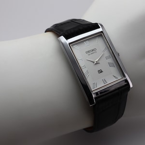 Könnte beinhalten: Eine rechteckige Seiko Quartz-Uhr mit silberfarbenem Gehäuse und weißem Zifferblatt mit römischen Ziffern. Die Uhr hat ein schwarzes Lederarmband. Die Uhr wird auf einer weißen Oberfläche präsentiert.