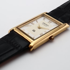 Puede incluir: Un reloj Seiko Quartz dorado con una esfera rectangular y una correa de cuero negro. La esfera es de color crema con n&uacute;meros romanos y el logotipo de Seiko. La correa es texturizada y el reloj est&aacute; en &aacute;ngulo.