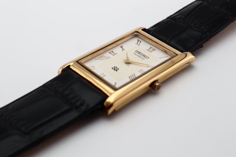 Puede incluir: Un reloj Seiko Quartz dorado con una esfera rectangular y una correa de cuero negro. La esfera es de color crema con n&uacute;meros romanos y el logotipo de Seiko. La correa es texturizada y el reloj est&aacute; en &aacute;ngulo.