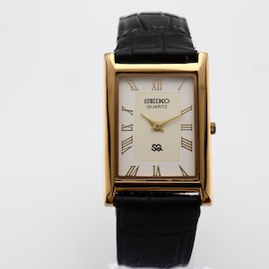 Puede incluir: Un reloj Seiko Quartz rectangular dorado con esfera color crema y correa de cuero negro. La esfera del reloj presenta n&uacute;meros romanos y agujas doradas. La correa es negra y texturizada.