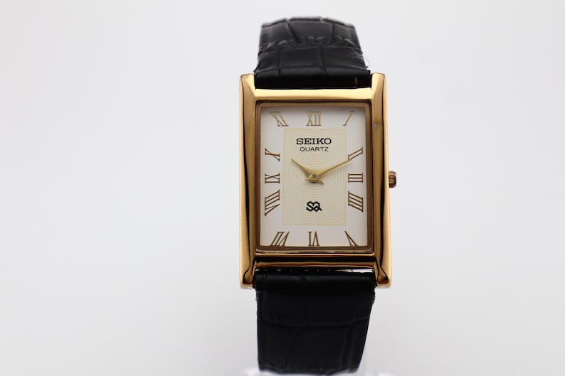 Puede incluir: Un reloj Seiko Quartz rectangular dorado con esfera color crema y correa de cuero negro. La esfera del reloj presenta n&uacute;meros romanos y agujas doradas. La correa es negra y texturizada.