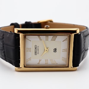 Puede incluir: Reloj Seiko Quartz dorado con esfera rectangular y correa de cuero negro. La esfera es blanca con n&uacute;meros romanos y agujas doradas. La correa tiene un patr&oacute;n de cocodrilo.