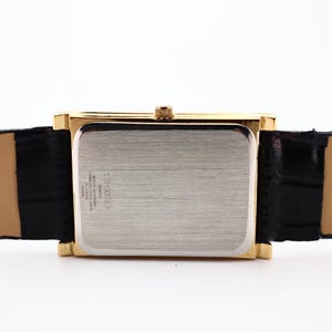 Puede incluir: Una caja de reloj rectangular dorada con una parte trasera plateada y una correa de cuero negra. La correa del reloj est&aacute; sujeta a la caja del reloj. El reloj tiene una corona dorada. La correa del reloj es negra.