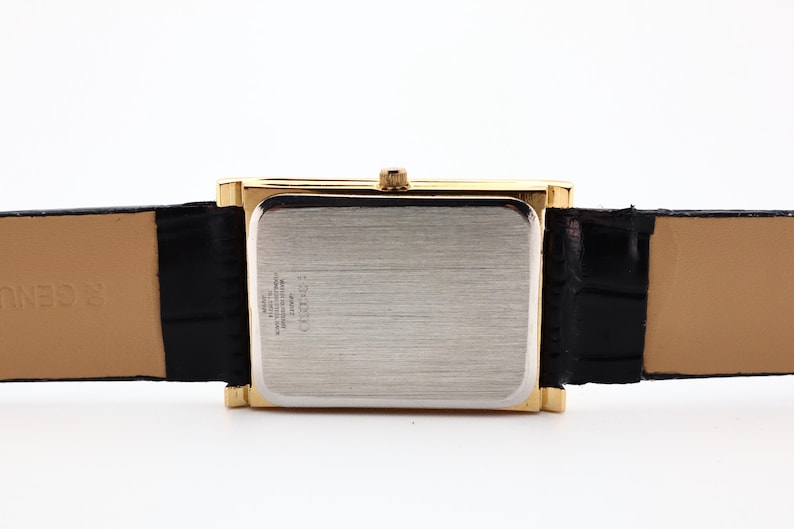 Puede incluir: Una caja de reloj rectangular dorada con una parte trasera plateada y una correa de cuero negra. La correa del reloj est&aacute; sujeta a la caja del reloj. El reloj tiene una corona dorada. La correa del reloj es negra.