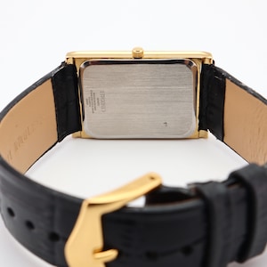 Puede incluir: Un reloj rectangular dorado con una correa de cuero negro. La esfera del reloj es plateada y la correa tiene una hebilla dorada. El reloj se ve desde la parte posterior, mostrando la textura de la correa y los detalles del reloj.