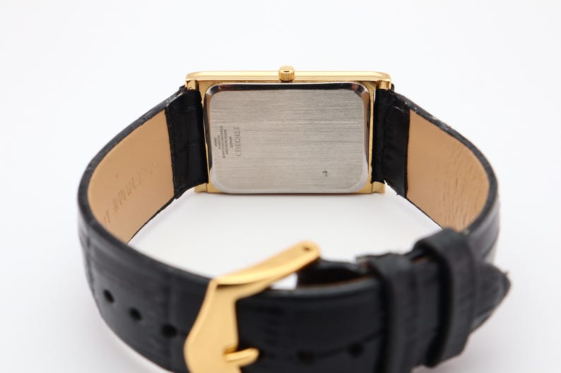 Puede incluir: Un reloj rectangular dorado con una correa de cuero negro. La esfera del reloj es plateada y la correa tiene una hebilla dorada. El reloj se ve desde la parte posterior, mostrando la textura de la correa y los detalles del reloj.