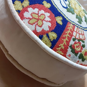 May include: Round, cream-colored cushion with a floral embroidered top. The embroidery features red, yellow, blue, and green flowers on a white background.